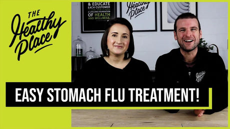 A smiling man and woman, both wearing black 'Find Your Healthy Place' branded apparel, sit together. A bold black and white text overlay reads 'EASY STOMACH FLU TREATMENT!' with 'The Healthy Place' logo on the left side.