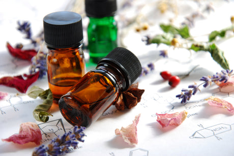 Small amber and green glass bottles of essential oils rest on a sheet of chemical formulas, surrounded by dried herbs, flowers, and spices.