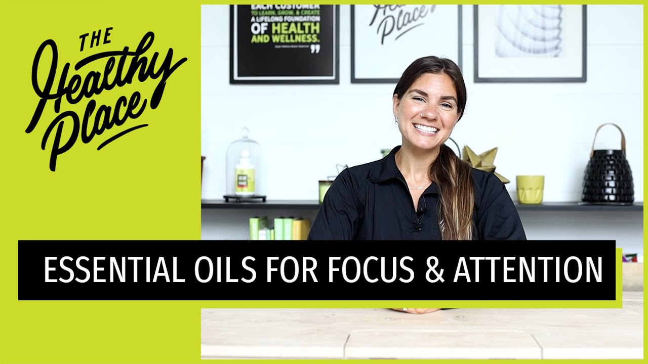 A video thumbnail from "The Healthy Place" featuring a smiling woman at a table with bold text reading "ESSENTIAL OILS FOR FOCUS & ATTENTION" and the brand’s logo on a lime green background.