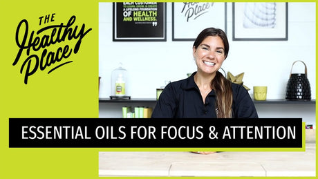 A video thumbnail from "The Healthy Place" featuring a smiling woman at a table with bold text reading "ESSENTIAL OILS FOR FOCUS & ATTENTION" and the brand’s logo on a lime green background.
