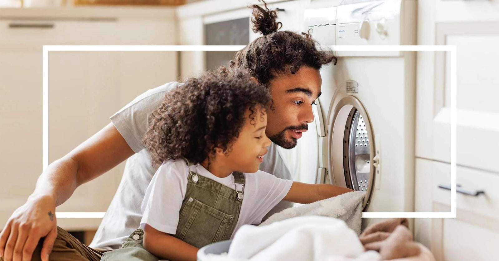 A father and child smile as they load laundry into a washing machine. A white rectangular overlay frames their faces.