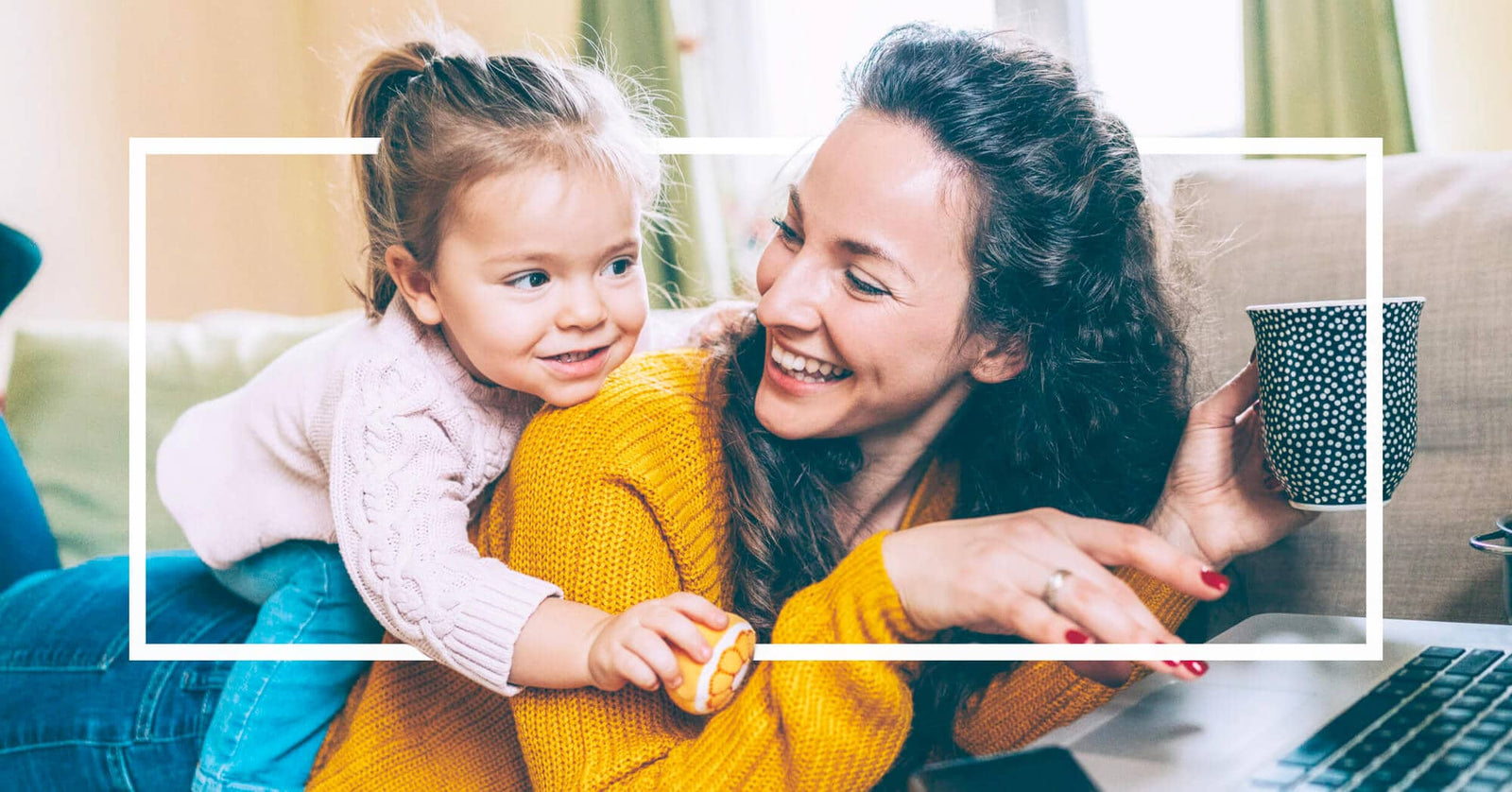 A smiling mother in a yellow sweater works on her laptop while her young daughter playfully climbs on her back.