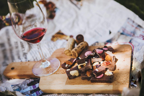A glass of red wine and a wooden cutting board filled with dark chocolate pieces topped with nuts, dried fruit, and pink candy shells.