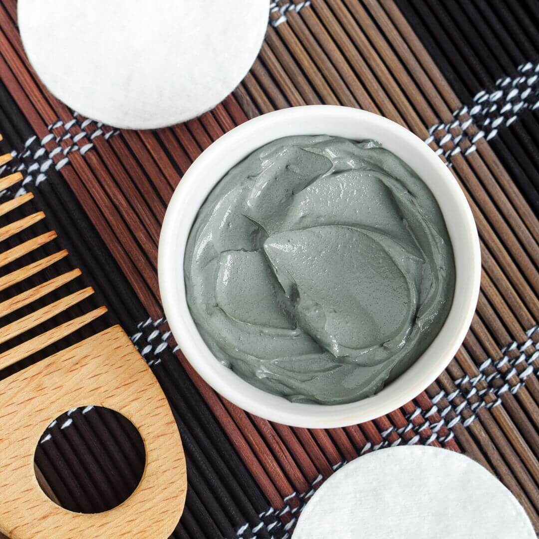 A small white bowl filled with a smooth, grayish-green clay mask sits on a woven bamboo mat. Surrounding it are a wooden comb with a finger hole and two round cotton pads, creating a spa-like, natural skincare setup.