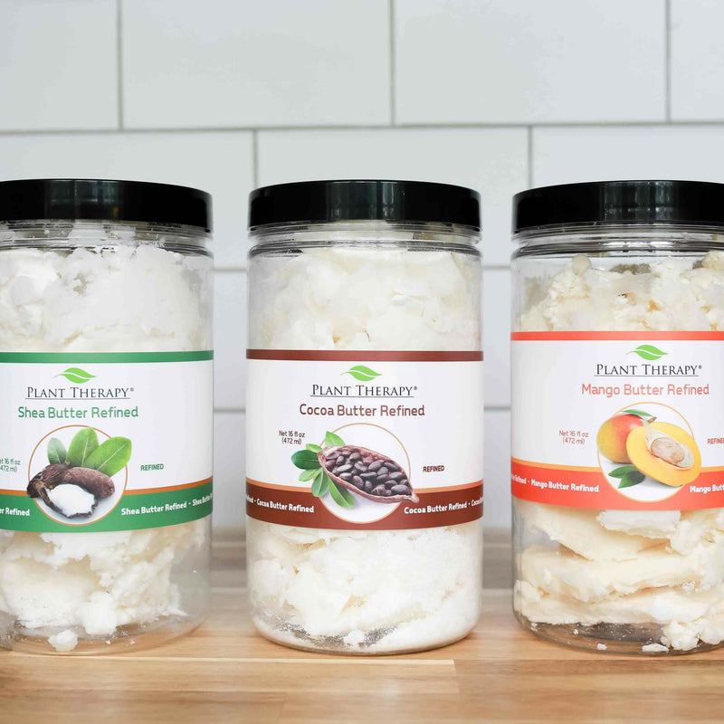 Three jars of refined body butters from Plant Therapy — Shea Butter, Cocoa Butter, and Mango Butter — are displayed on a wooden surface against a white tile background. Each jar contains labels featuring images of the respective natural ingredients.