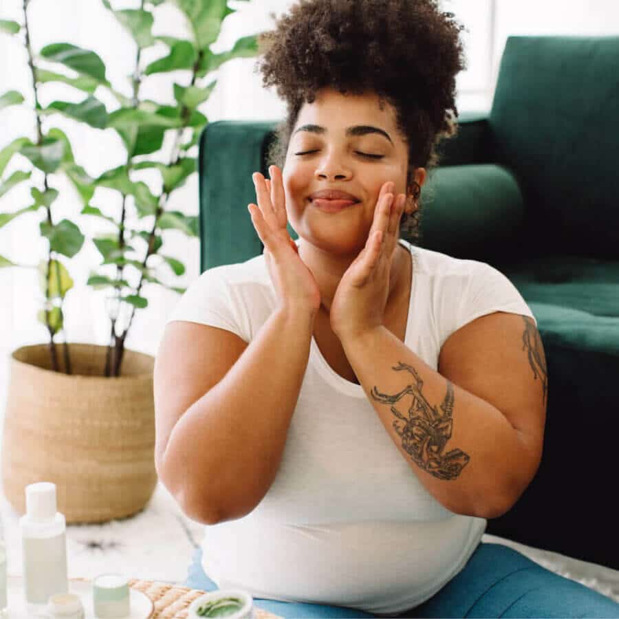 A woman with curly hair and tattoos smiles with her eyes closed while gently touching her face. She sits in a cozy space with a green couch and potted plants, surrounded by skincare products, suggesting a self-care routine.