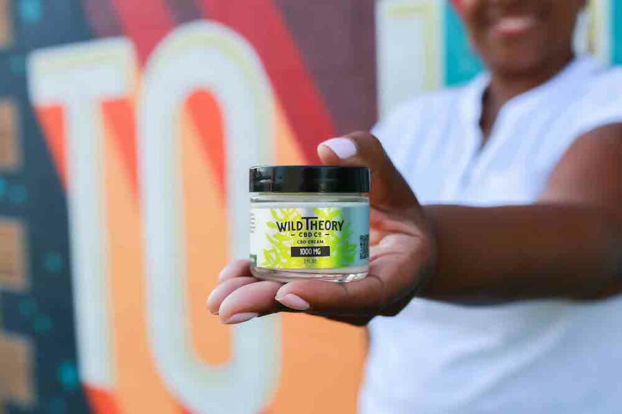 A smiling woman in a white shirt holds out a jar of Wild Theory CBD Co. CBD cream (1000 MG) toward the camera. The background features a colorful mural, creating a vibrant and energetic setting.