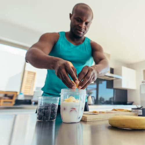 A muscular man in a teal tank top prepares a post-workout smoothie in a modern kitchen. He is adding fruit slices to a blender cup filled with milk and berries, with a container of blueberries and a banana on the counter.