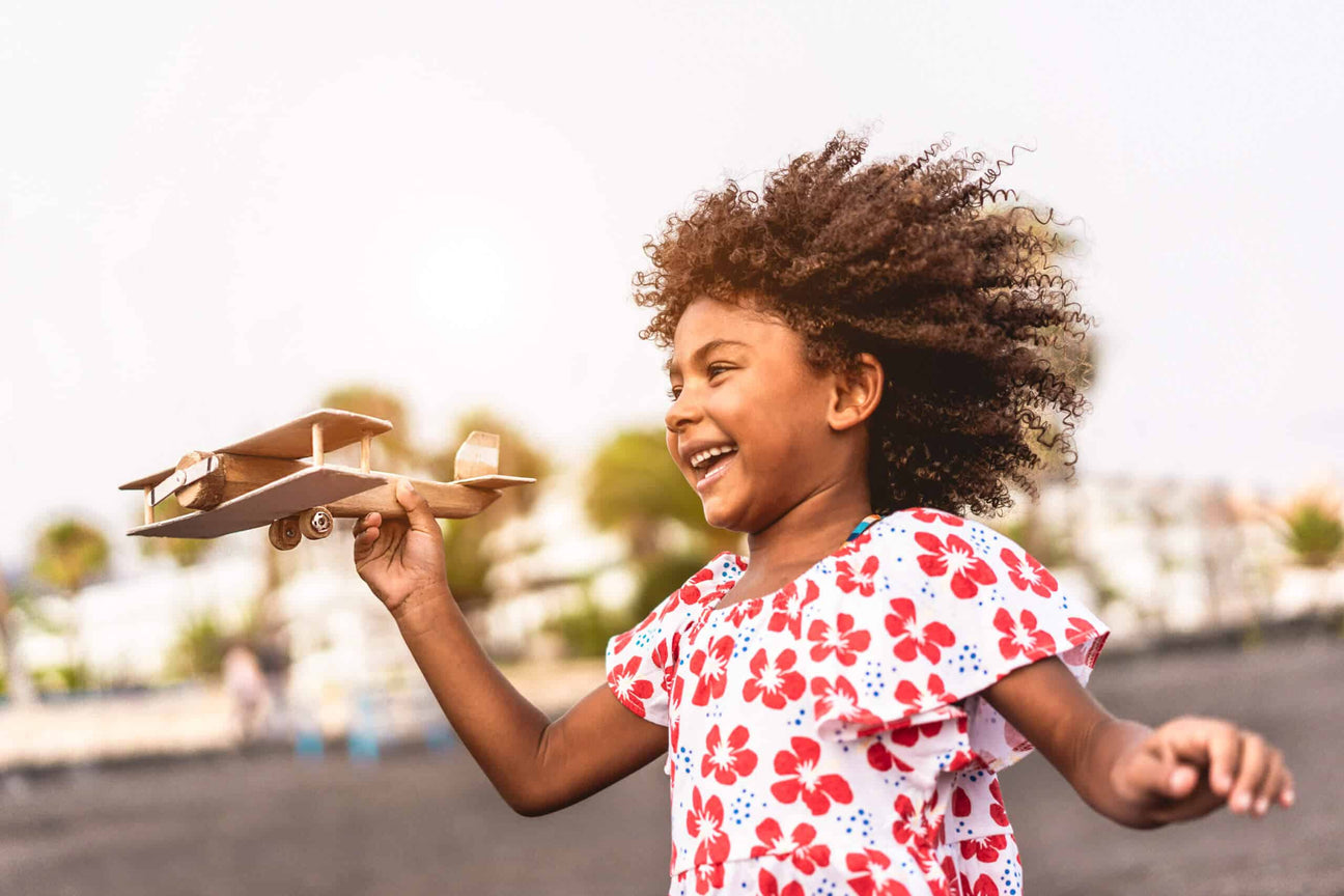 A joyful young girl with curly hair plays outdoors, holding a wooden toy airplane in one hand. She wears a white and red floral dress, her face lit up with laughter as she moves energetically.
