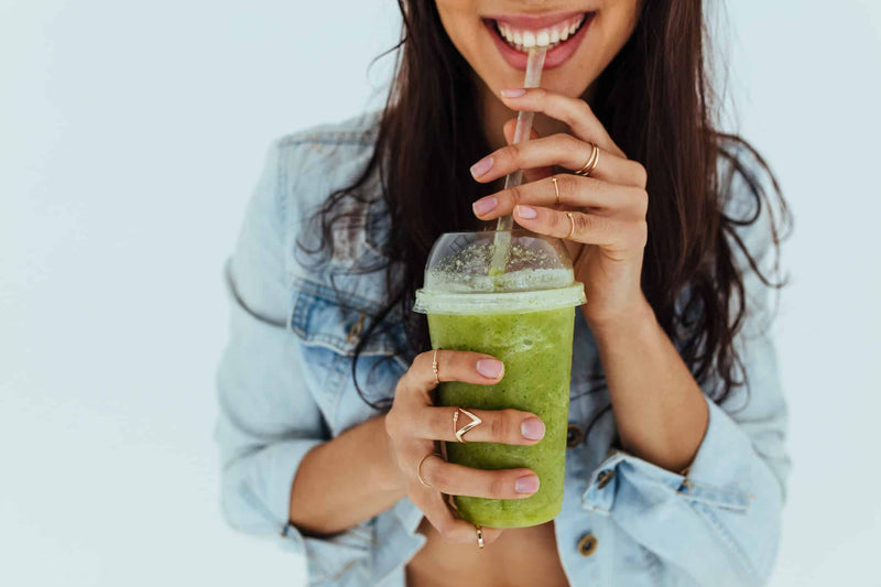 A smiling woman in a denim jacket sips a vibrant green smoothie from a clear plastic cup with a straw. Her hands, adorned with delicate gold rings, gently hold the cup.
