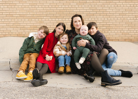 Becki OBrien, owner of The Healthy Place, sitting on the concrete with all of her children gathered around her. They're all smiling and cuddling closely together in front of a brick wall.