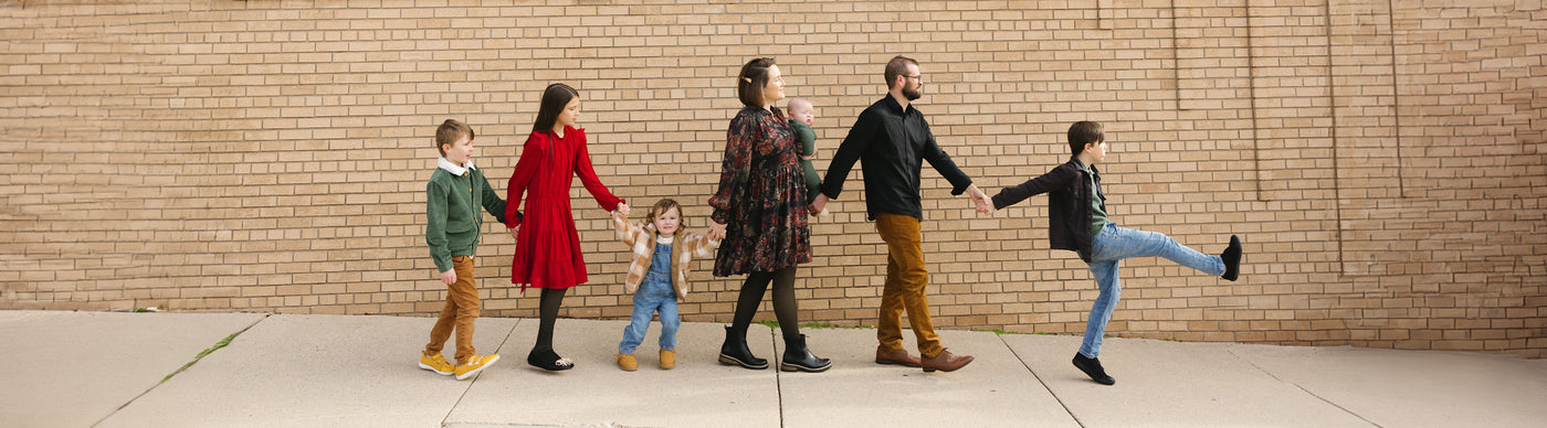The OBrien family walking in a row in front of a brick wall. A young boy leads the line, kicking his leg up, Tim behind, and Becki holding their new baby boy. A toddler smiles at the camera, followed by his big sister holding his hand. Another young boy brings up the rear with a smile.