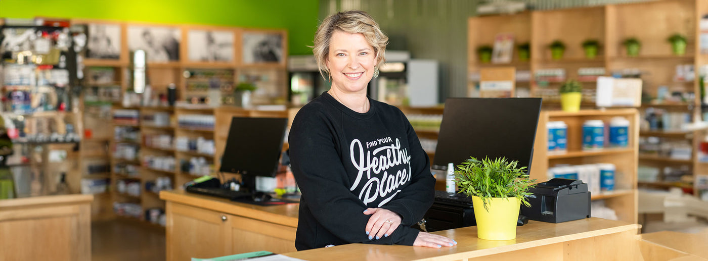 Kenzie, a Wellness Consultant of The Healthy Place, smiling sweetly at the register of the Fitchburg store. Shelves full of supplements are blurred behind her.