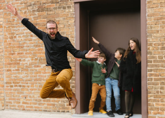 Tim OBrien, owner of The Healthy Place, leaping in the air with a wide smile and his arms outstretched. Behind him are several of his children in a doorway posing and laughing.