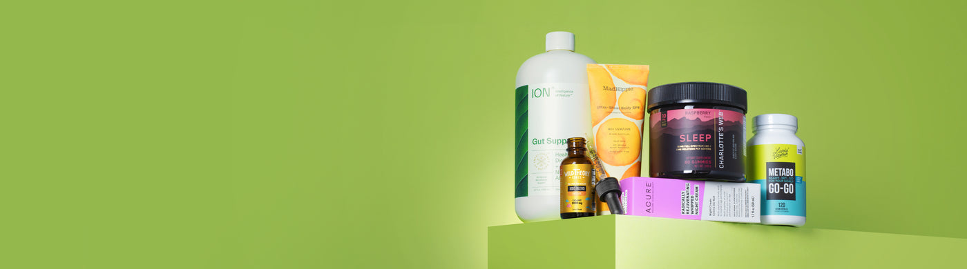 A group of wellness products arranged on a green platform against a light green background. Items include a large bottle of ION Gut Support, Mad Hippie Ultra-Sheer Body SPF, Wild Theory CBD oil, Charlotte's Web Sleep gummies, ACURE night cream, and a bottle of Metabo Go-Go capsules. The arrangement highlights a variety of supplements and skincare for health and self-care.