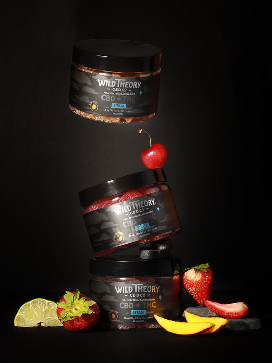 Three stacked jars of Wild Theory CBD + THC gummies are artistically arranged against a dark background, with a cherry balancing between two jars. Fresh fruit, including strawberries, mango slices, and an apple, surround the base, highlighting the fruity flavors.