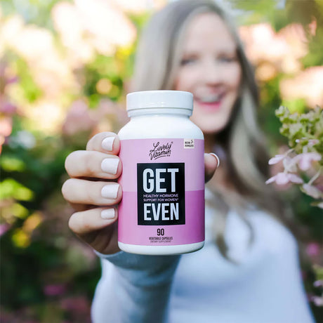 A smiling woman in soft focus holds a white bottle of Lively Vitamin Co.'s “Get Even” supplement toward the camera; the label is pink and black with bold text stating “Healthy Hormone Support for Women – 90 capsules,” framed by a vibrant outdoor setting with flowers and greenery.