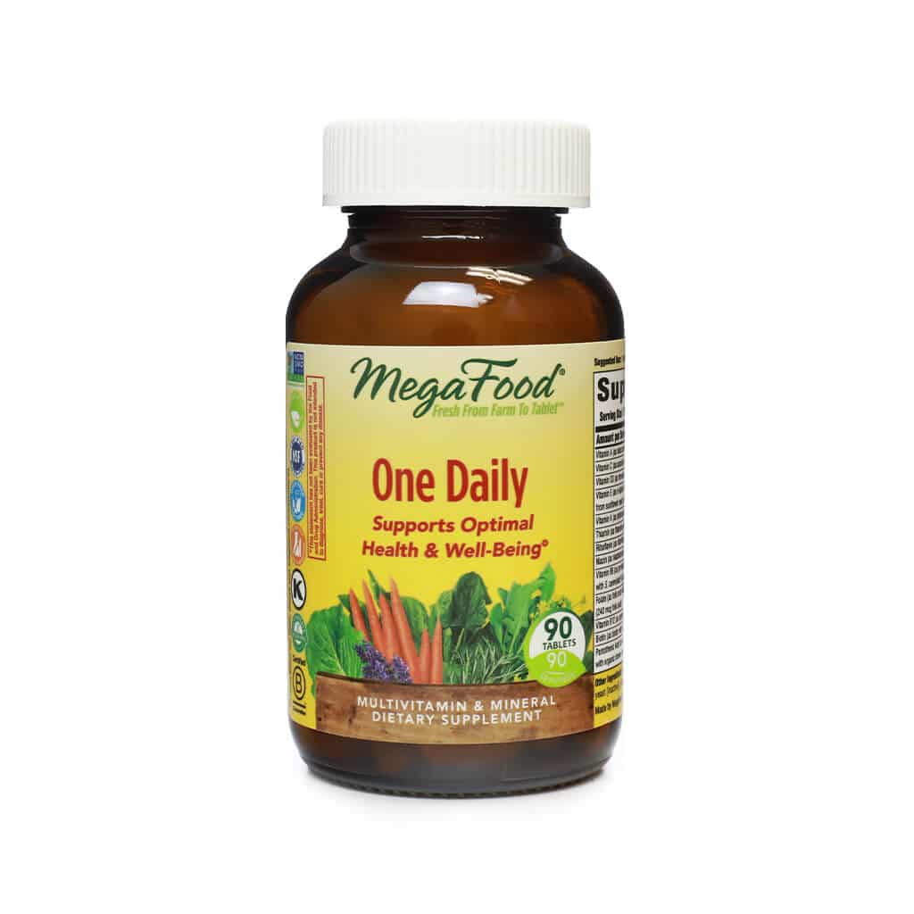 A brown glass bottle labeled "MegaFood One Daily" multivitamin, containing 90 tablets. The label is yellow with green accents and features images of carrots, leafy greens, and lavender, emphasizing support for optimal health and well-being.