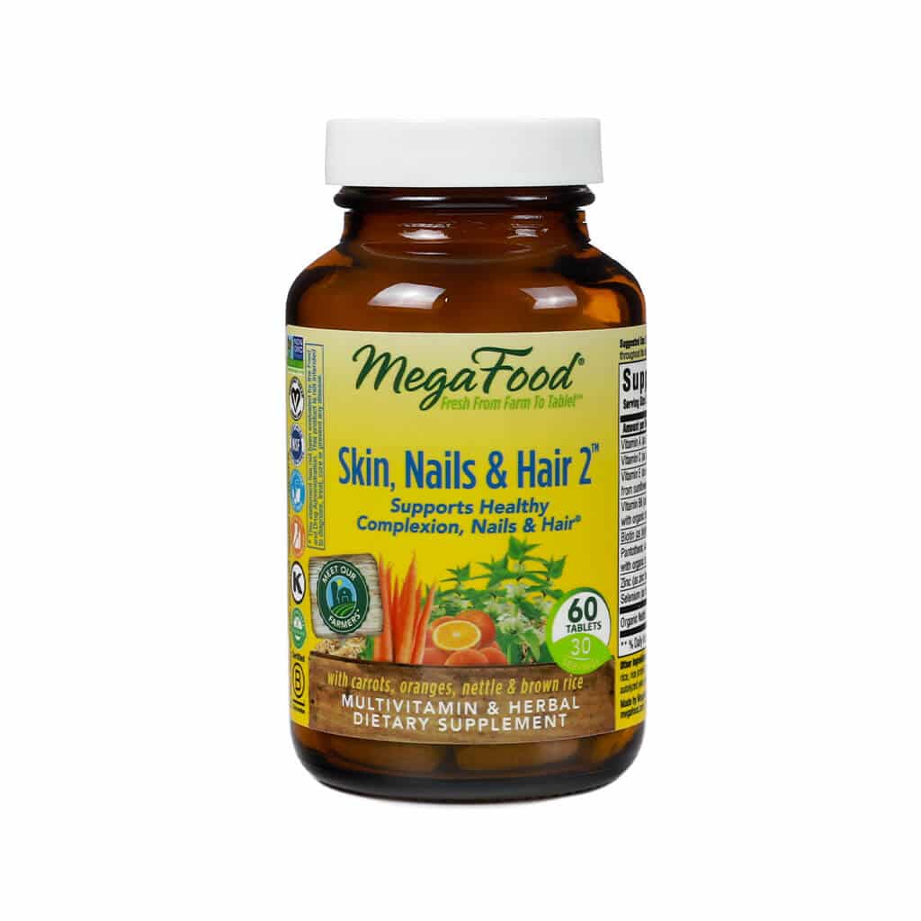 A brown bottle of MegaFood Skin, Nails & Hair 2 supplement, featuring images of carrots, oranges, and nettle. The label highlights its support for healthy complexion, nails, and hair, with 60 tablets per bottle, and includes vitamins and herbs like biotin and brown rice.
