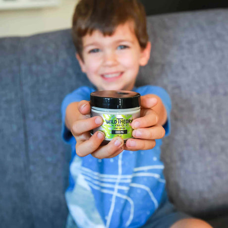 A young child with a big smile holds a jar of Wild Theory CBD Cream 1000mg toward the camera, with the product label clearly visible. The child is sitting on a gray couch, wearing a blue shirt topped with an intersellar design.