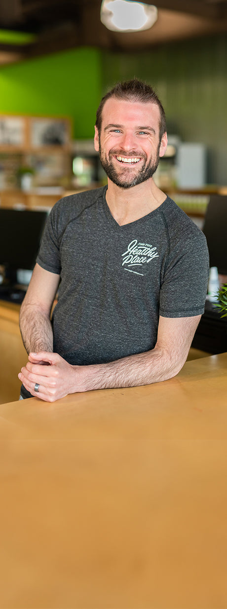 Tim O’Brien, owner of The Healthy Place, stands behind a wooden counter smiling warmly in a wellness store setting. He’s wearing a dark gray T-shirt with “Find Your Healthy Place” printed on it, with shelves and green walls visible in the background.