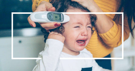 A crying young child with curly brown hair and flushed cheeks is held by an adult wearing a mustard-yellow sweater. The adult is using a digital ear thermometer to check the child's temperature. A white rectangular frame overlays the image.