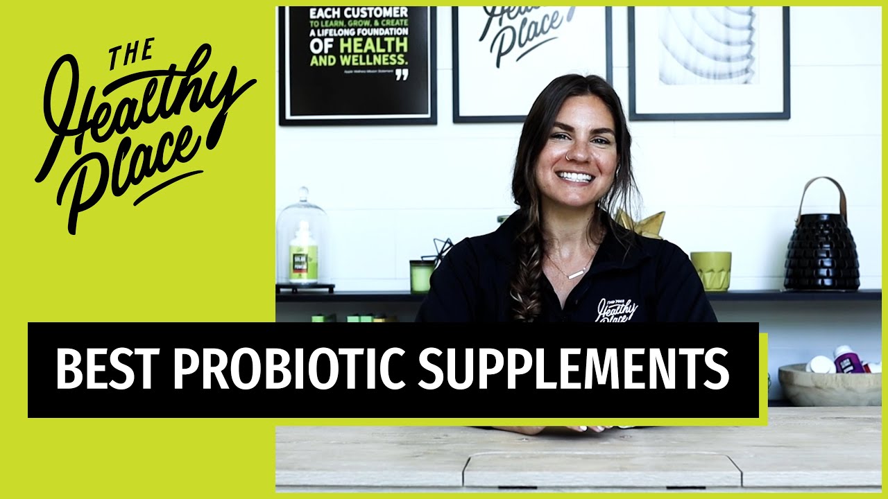A smiling woman with long brown hair sits at a wooden table. old text overlay reads 'BEST PROBIOTIC SUPPLEMENTS' on a black and green banner, with 'The Healthy Place' logo prominently displayed on the left side.