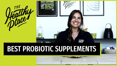 A smiling woman with long brown hair sits at a wooden table. old text overlay reads 'BEST PROBIOTIC SUPPLEMENTS' on a black and green banner, with 'The Healthy Place' logo prominently displayed on the left side.