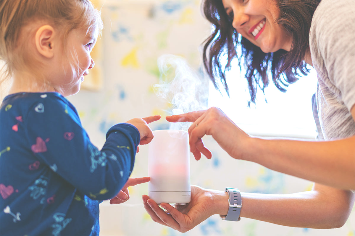 A  smiling mother and her young daughter playfully touch the mist coming from a small white essential oil diffuser. 