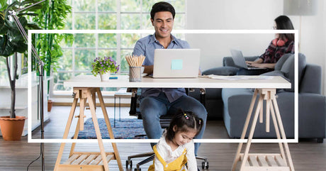 A man works at his desk while a young girl plays on the floor, and a woman uses a laptop in the background.