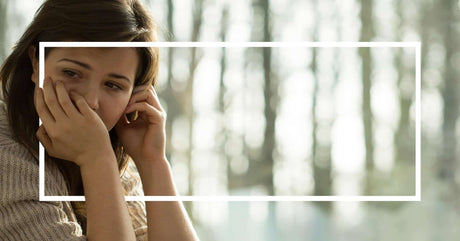 A young woman sits outdoors, resting her head on her hand with a worried expression. A white rectangular border frames the image.