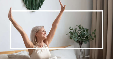 A smiling older woman with short gray hair stretches her arms wide while sitting up in bed. She appears well-rested and content, with soft morning light illuminating her cozy bedroom.