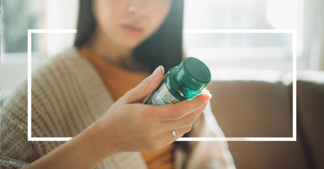 A close-up of a woman holding a green supplement bottle, reading the label carefully while sitting in a softly lit room.