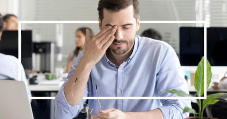 A man in a blue shirt rubs his eyes at his desk, holding glasses, with a white rectangular border framing the image.