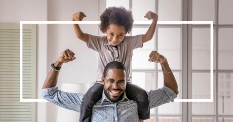 Smiling man with a child on his shoulders, both flexing their arms in a playful strong pose.