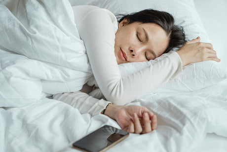 A woman in a white long-sleeve shirt sleeps soundly on a white bed, hugging a pillow. Her hand rests near a smartphone, suggesting she may have fallen asleep after using it.