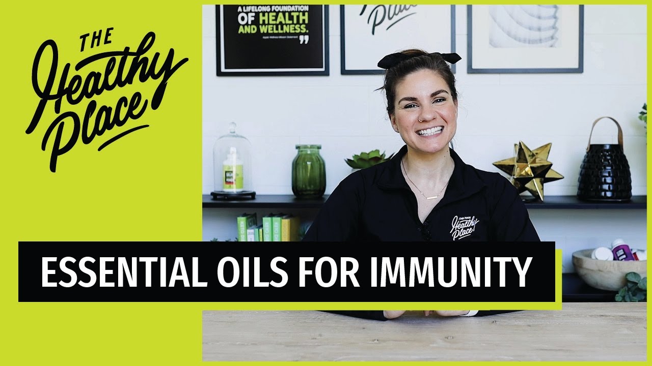 Smiling woman in a 'Find Your Healthy Place' jacket sits at a table with a wellness-themed background. Bold text overlay reads 'ESSENTIAL OILS FOR STRESS.'