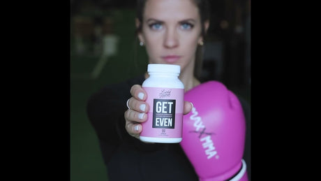 A woman in a black outfit holds 'GET EVEN' hormone support supplements, wearing a pink boxing glove, symbolizing strength and wellness.