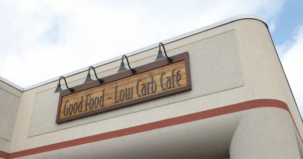 Signage reading "Good Food - Low Carb Café" mounted on the exterior wall of a building with decorative lighting above.