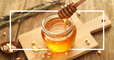 A jar of golden honey with a wooden dipper, surrounded by pine nuts and rustic kitchen elements on a wooden table.