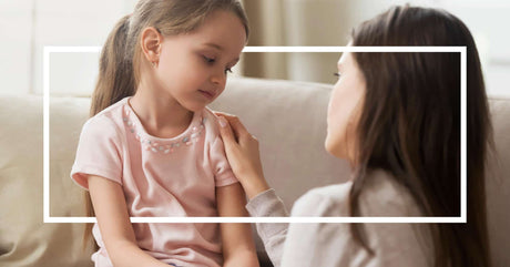 A woman gently comforts a sad young girl sitting on a couch. 