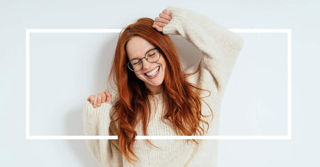 A joyful red-haired woman in glasses stretches her arms while smiling.