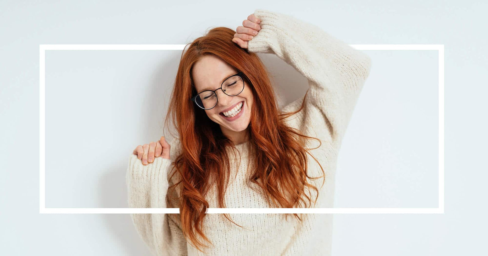 A joyful red-haired woman in glasses stretches her arms while smiling.