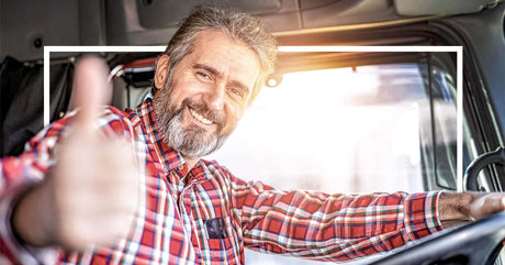 A smiling truck driver in a red plaid shirt gives a thumbs-up inside his vehicle, with sunlight streaming through the window.