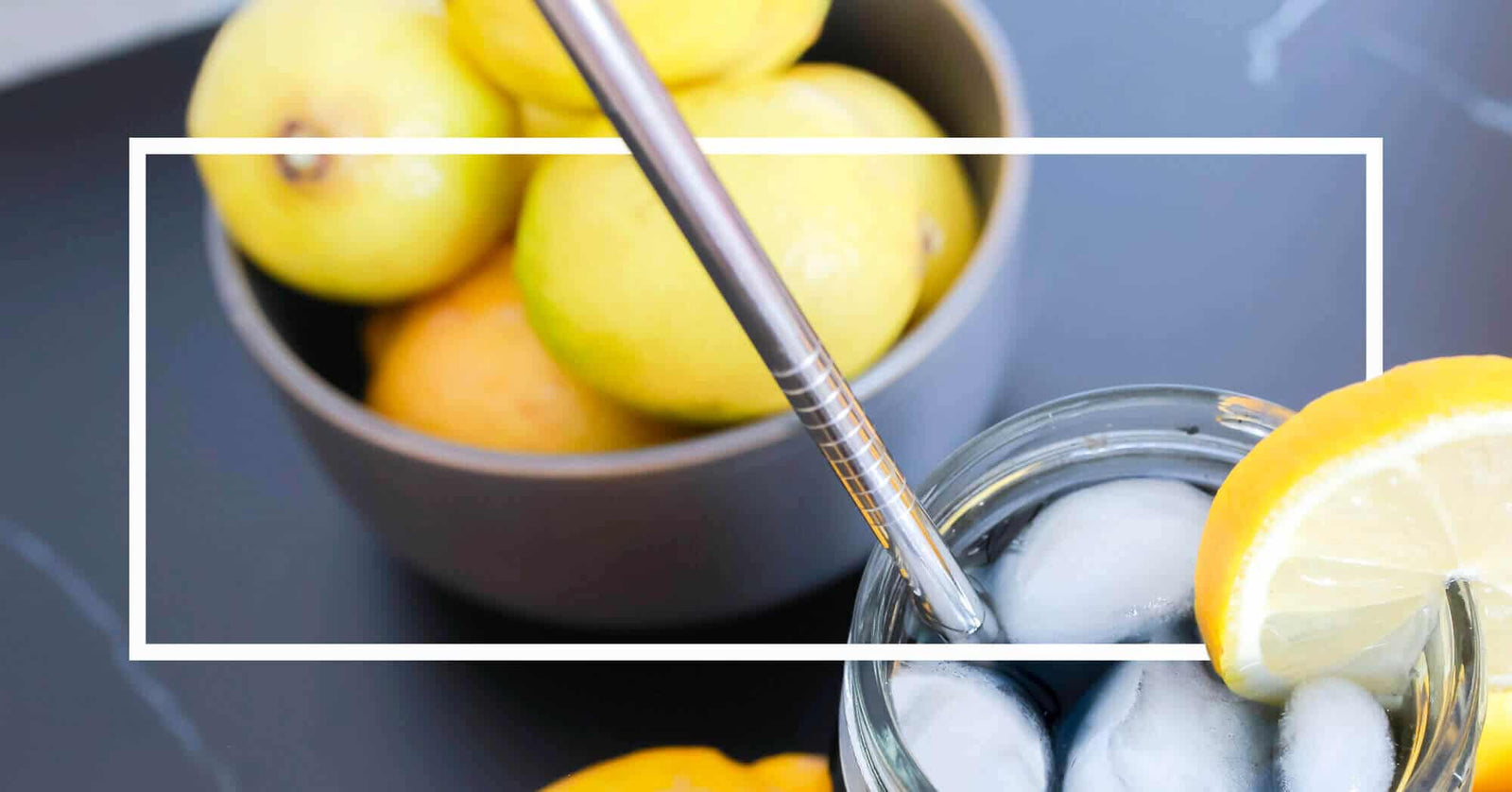 A glass of iced lemon water with a metal straw next to a bowl of fresh citrus fruits.