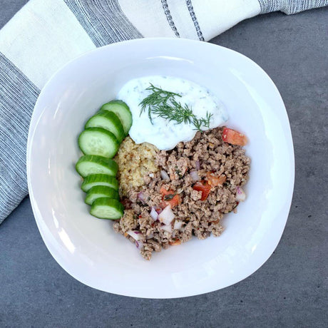 A white bowl containing a Mediterranean-inspired dish with sliced cucumbers, seasoned ground beef, diced tomatoes and onions, quinoa, and a dollop of tzatziki sauce garnished with fresh dill.