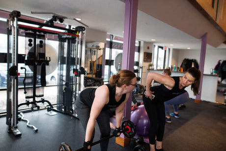 A personal trainer in black athletic wear coaches a woman lifting a barbell in a well-lit gym, demonstrating proper form with a hand on her lower back.