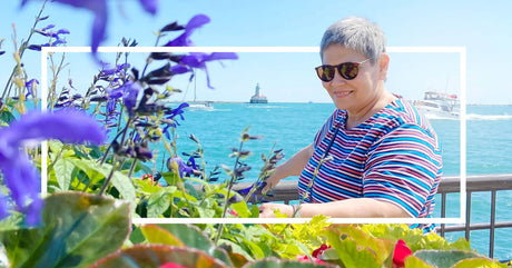 A smiling woman in sunglasses and a striped shirt leans on a railing by the water, with vibrant flowers in the foreground and a lighthouse in the distance.