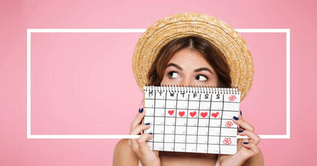 A young woman wearing a straw hat holds up a spiral-bound calendar in front of her face, revealing only her expressive eyes. The calendar displays a month layout with days marked, and several dates are highlighted with red heart symbols.