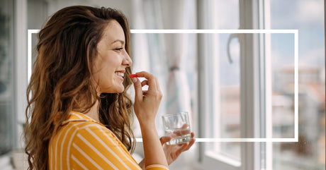 A smiling woman in a yellow striped shirt holds a red capsule and a glass of water by a bright window.